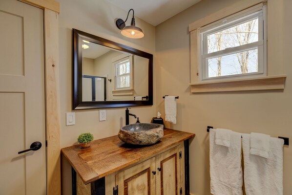 A bathroom with a wooden vanity, a stone sink, a large mirror, a towel rack, and a window with a view of trees.