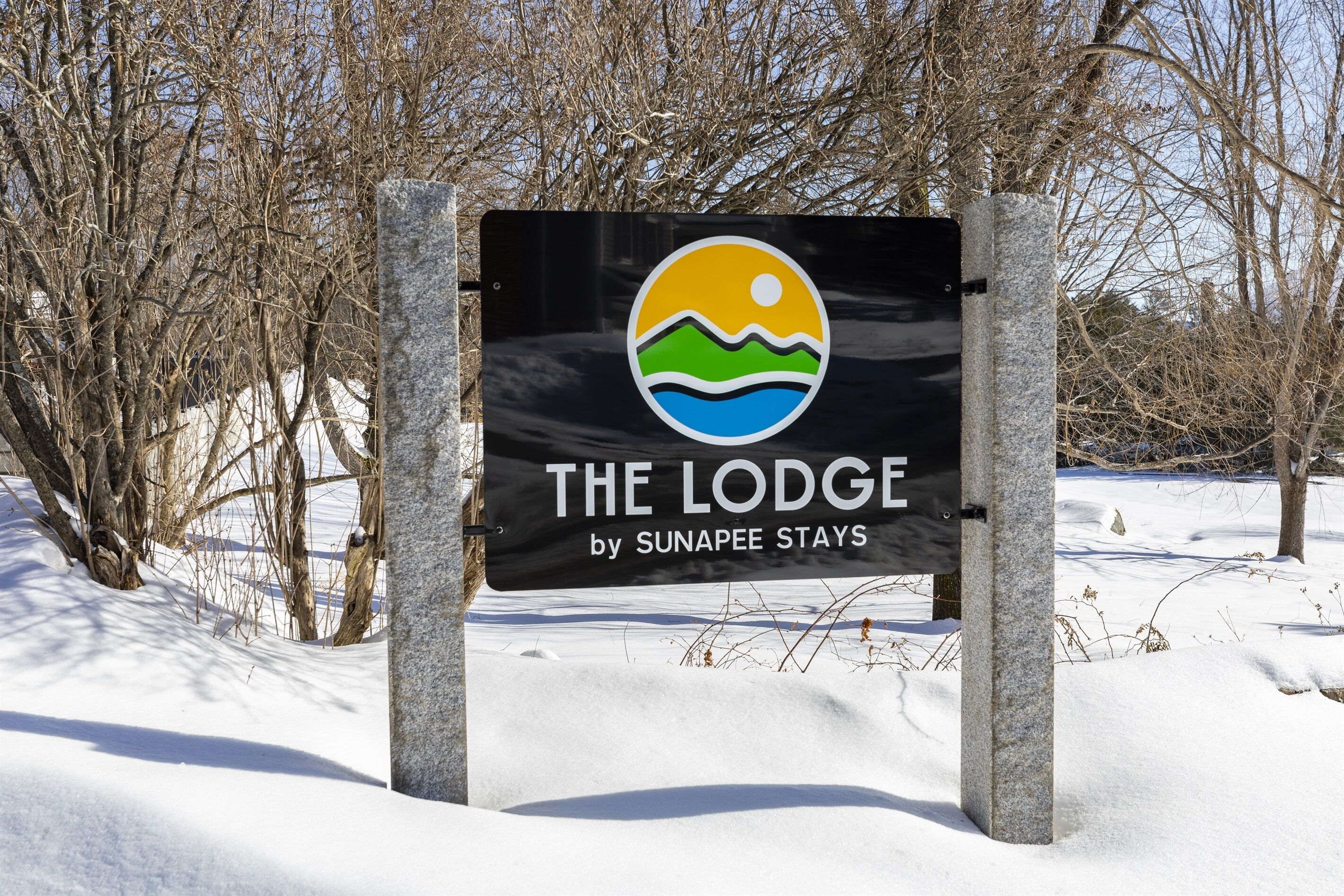 A sign for 'The Lodge by Sunapee Stays' with a logo featuring a sun, waves, and a mountain.
