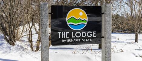 A sign for 'The Lodge by Sunapee Stays' with a logo featuring a sun, waves, and a mountain.