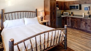 A rustic bedroom with a wooden bed, a nightstand with a lamp, and a kitchenette with wooden cabinets and modern appliances.