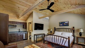 Un dormitorio con techo de madera, una cama, un televisor y una zona de cocina con armarios de madera y electrodomésticos.