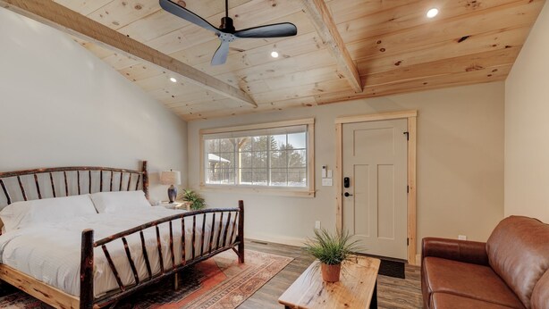 Un dormitorio con techo de madera, cama con cabecera de madera, ventana, puerta, una mesita con una planta y un sofá de cuero marrón.