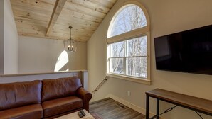 Un salon chaleureux avec un plafond en bois, un canapé en cuir, un téléviseur fixé au mur et une grande fenêtre donnant sur la forêt.