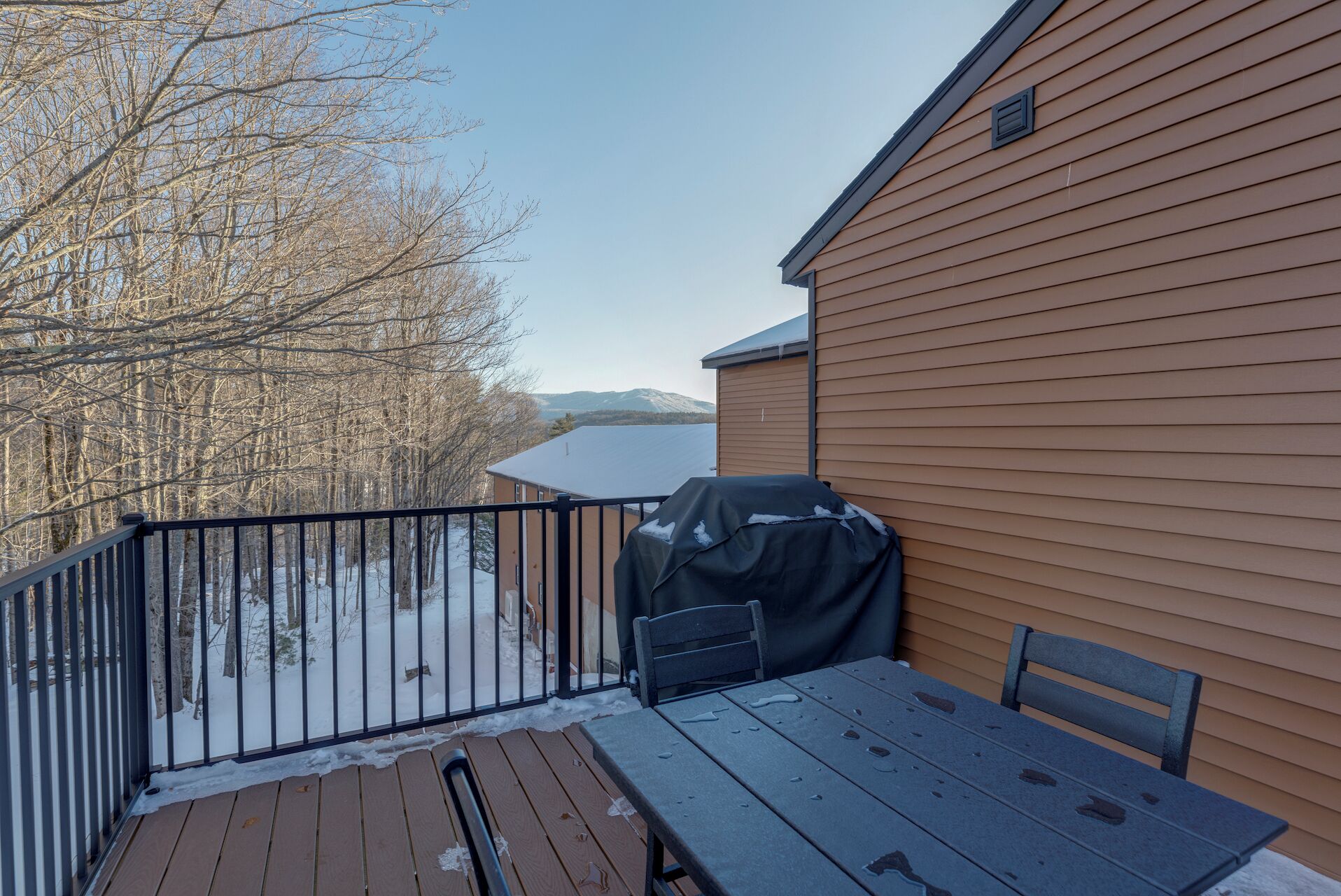 A deck with a table and chairs, a barbecue grill, and a view of a snowy landscape with trees and mountains.