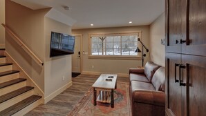 Ein Wohnzimmer mit Treppe, einem an der Wand befestigten Flachbildfernseher, einem Ledersofa, einem Couchtisch und einem Fenster mit Blick auf Bäume.