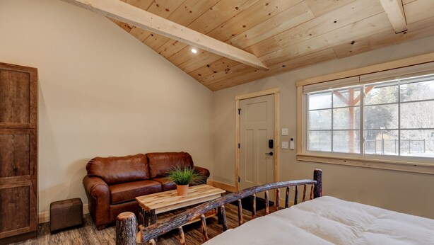 Una habitación acogedora con techo de madera, un sofá de cuero marrón, un banco de madera, una cama blanca y una ventana con vistas a árboles.