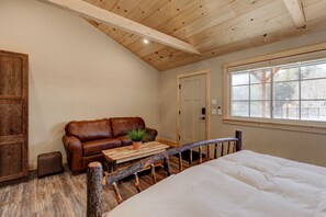 Una habitación acogedora con techo de madera, un sofá de cuero marrón, un banco de madera, una cama blanca y una ventana con vistas a árboles.