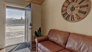 Un salon avec un canapé en cuir marron, une grande horloge en bois et une porte donnant sur un espace extérieur enneigé.