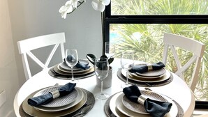 Une table de salle à manger ronde est dressée avec des chaises blanches, des assiettes, des couverts et des serviettes. Une orchidée blanche est placée dans un vase sur la table.