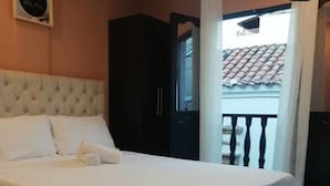 Un lit bien fait, avec du linge de lit blanc, une tête de lit sombre et une vue sur un balcon doté d’un toit en tuiles.