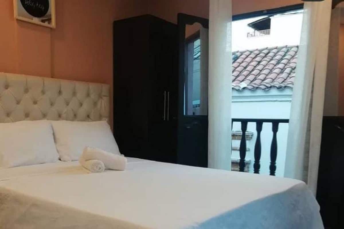 Un lit bien fait, avec du linge de lit blanc, une tête de lit sombre et une vue sur un balcon doté d’un toit en tuiles.