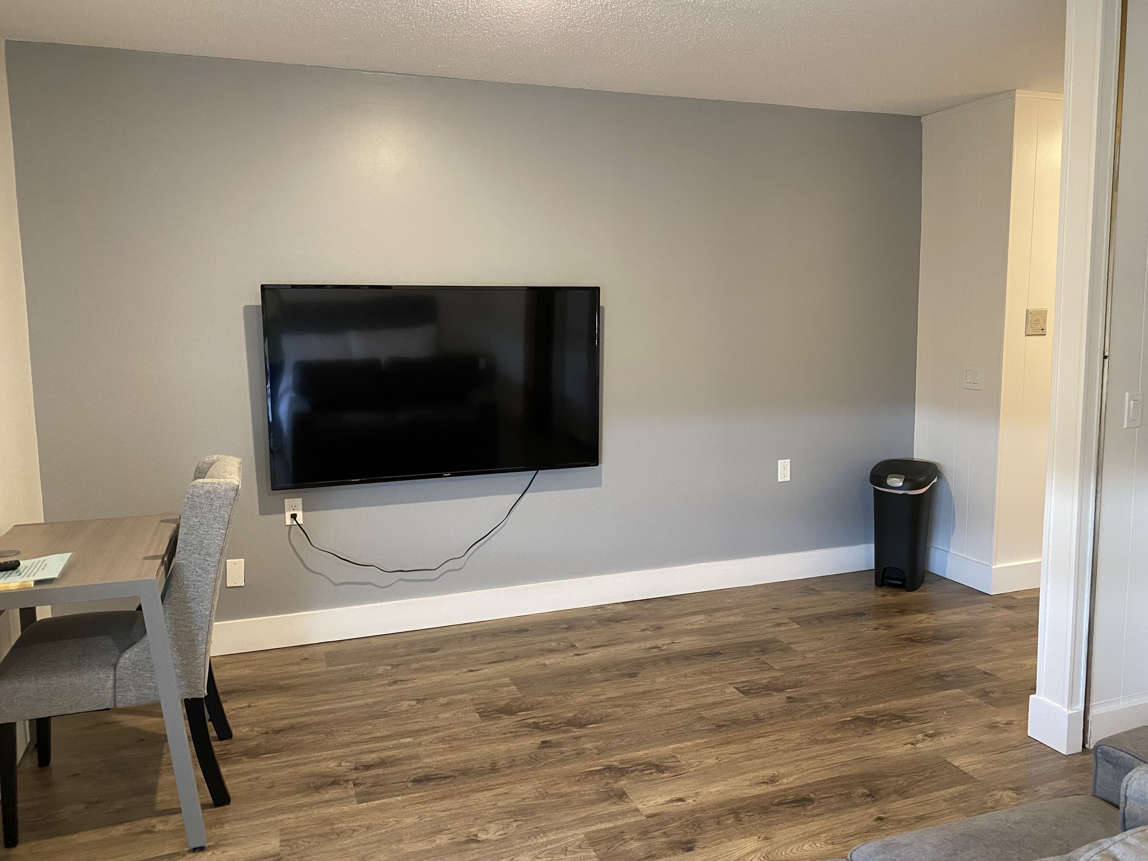 A modern living room with a flat-screen TV mounted on the wall, a dining table with chairs, and a trash can.