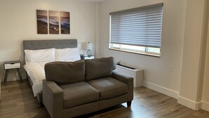 Ein Schlafzimmer mit einem Bett, einer grauen Kopfstütze, einem grauen Sofa, einem Nachttisch, einer Lampe und einem Fenster mit Jalousien.