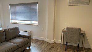 Ein Wohnzimmer mit grauem Sofa, einem kleinen Tisch und einem Sessel. Ein Fenster mit Jalousien, ein Wandbild und ein Holzboden sind ebenfalls zu sehen.