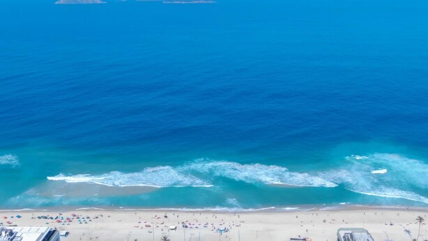 Uma praia com águas azuis cristalinas, ondas e uma faixa de areia com guarda-sóis e pessoas.