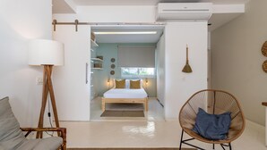 Quarto moderno com cama, uma cadeira, uma luminária e área de cozinha visível por uma porta aberta.