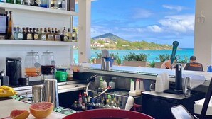 A bar area with a view of a beach and ocean, shelves with various liquor bottles, a red counter, and a large glass with a logo.