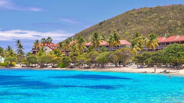 A beachfront hotel with multiple red-roofed buildings, palm trees, and clear blue water.