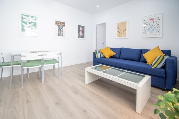 Una sala de estar moderna con un sofá azul, sillas verdes, una mesa de centro de vidrio y obras de arte en las paredes.