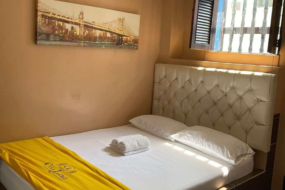 Ein Hotelzimmer mit Bett, Kopfteil, Fenster mit Fensterläden und einem Bild an der Wand.