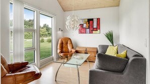 Uma sala de estar moderna com uma mesa de centro de vidro, poltronas de couro, um sofá cinza e uma grande janela com vista para uma paisagem verdejante.