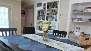 A dining area with a long table, chairs, a chandelier, a bookshelf, and a small cabinet.