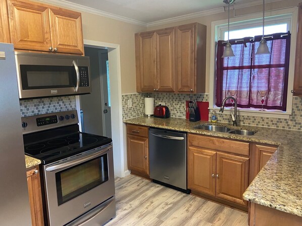A kitchen with wooden cabinets, a stainless steel microwave, oven, and dishwasher, a granite countertop, and a window with curtains.