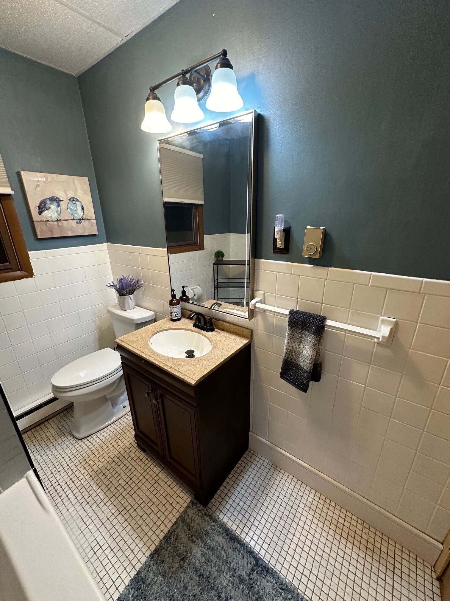 A bathroom with a dark blue wall, white tiled floor, a sink with a beige countertop, a mirror with three lights, a toilet, and a towel rack.