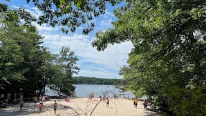 A beach with people sunbathing and swimming, surrounded by trees and a lake.