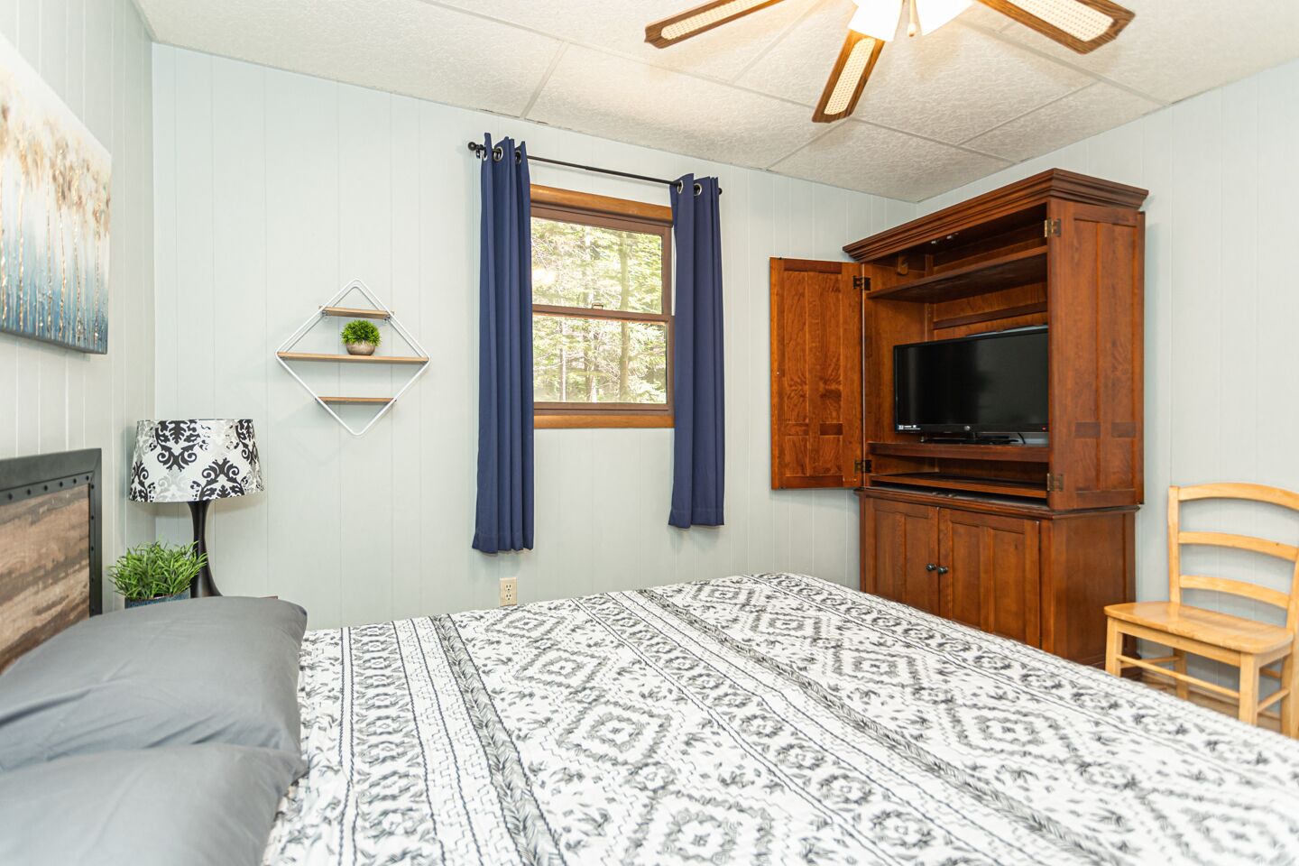 A bedroom with a bed, a TV, a wooden cabinet, a chair, and a window with curtains.