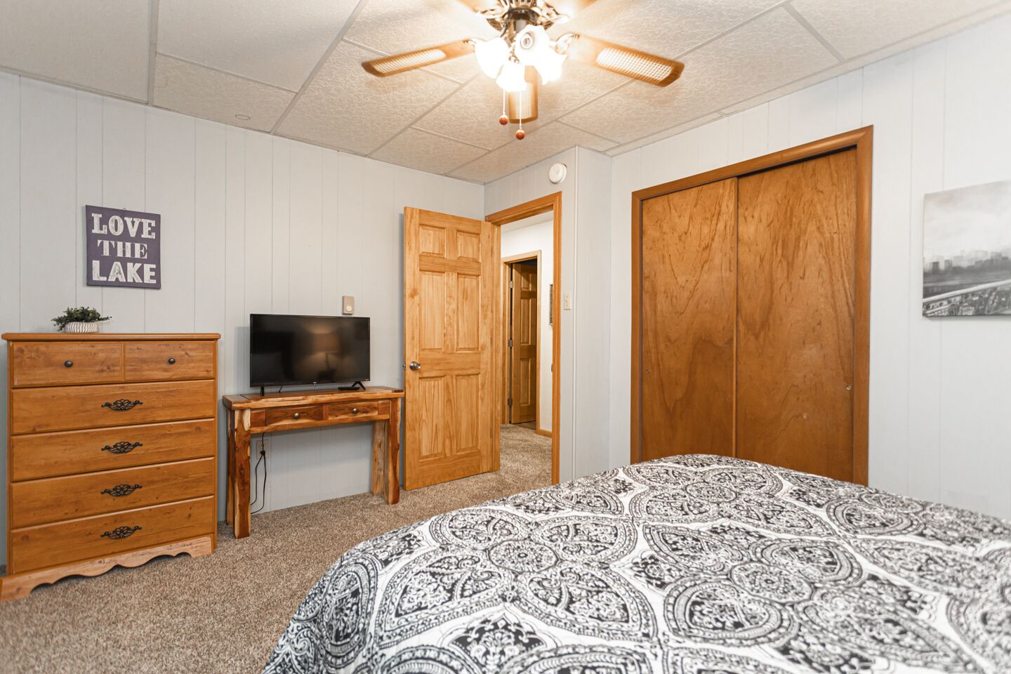 A bedroom with a bed, a dresser, a TV on a wooden desk, a ceiling fan, and a door leading to another room.