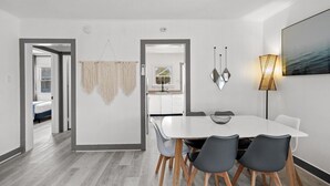 Een moderne eethoek met een witte tafel en grijze stoelen, een hangende macramé wanddecoratie, en een vloerlamp.