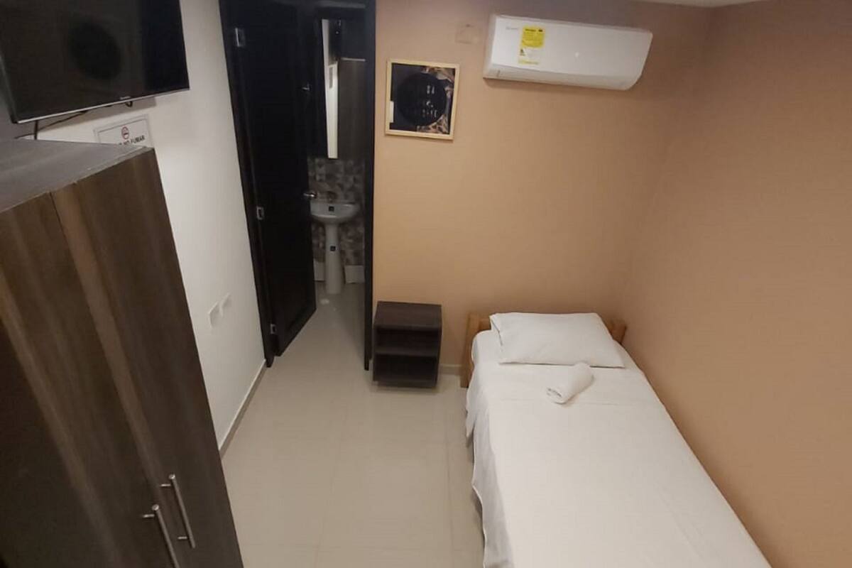 A small, well-lit room with a bed, a wardrobe, a sink, and a television.
