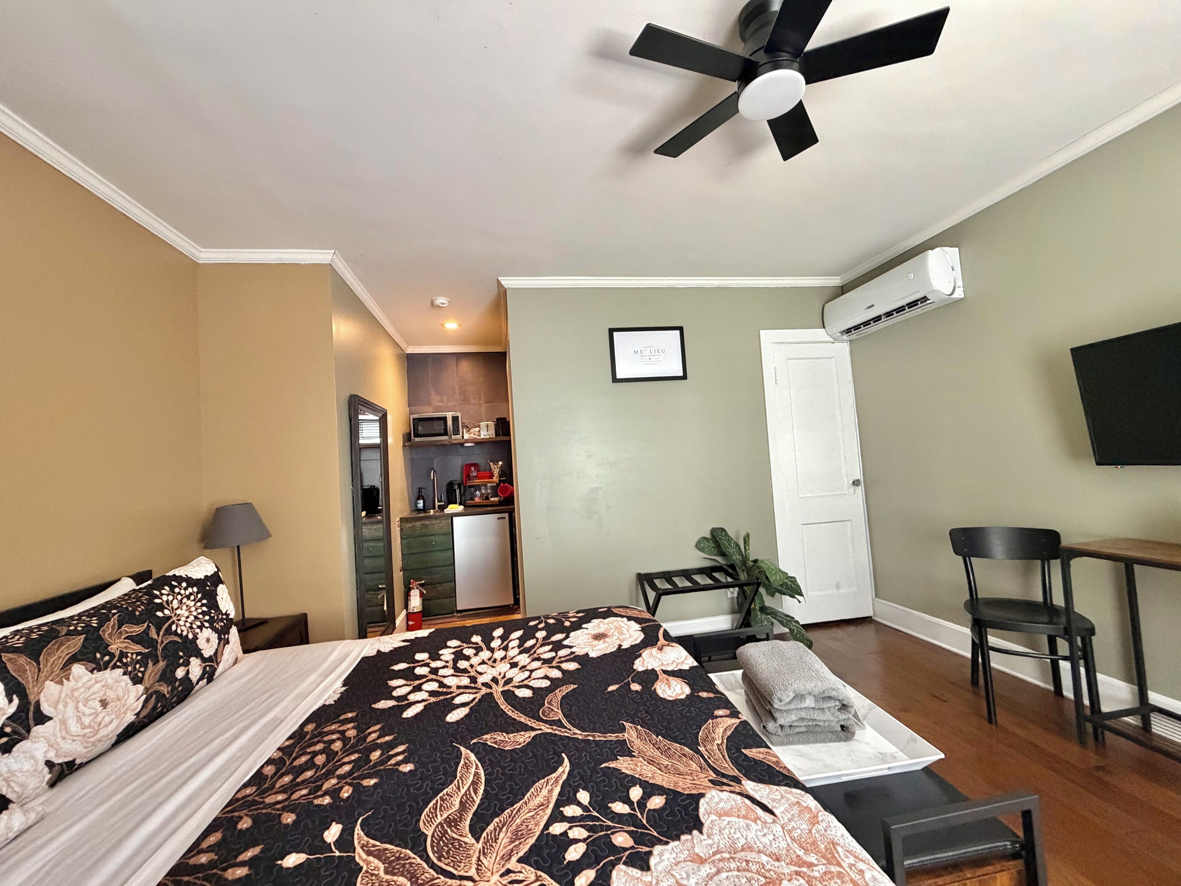 A bedroom with a bed, a desk, a chair, a ceiling fan, and a kitchenette in the background.