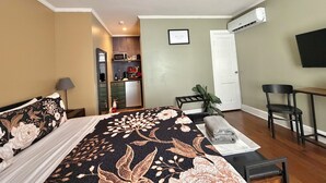 A bedroom with a bed, a desk, a chair, a ceiling fan, and a kitchenette in the background.