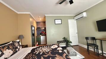 A bedroom with a bed, a desk, a chair, a ceiling fan, and a kitchenette in the background.
