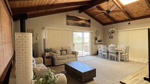 A spacious living area with a high ceiling, wooden beams, a ceiling fan, a large window, and a dining area with a table and chairs.