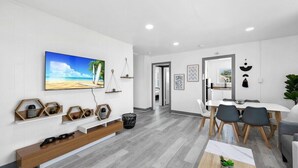 A modern living room with a flat-screen TV, wooden shelves, a dining area with a table and chairs, and a hallway leading to another room.