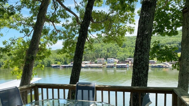 A glass table and chairs on a wooden deck overlooking a lake with boats and buildings in the background.