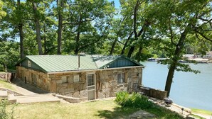 A stone cabin with a green roof by a lake, featuring a patio and a fire pit.