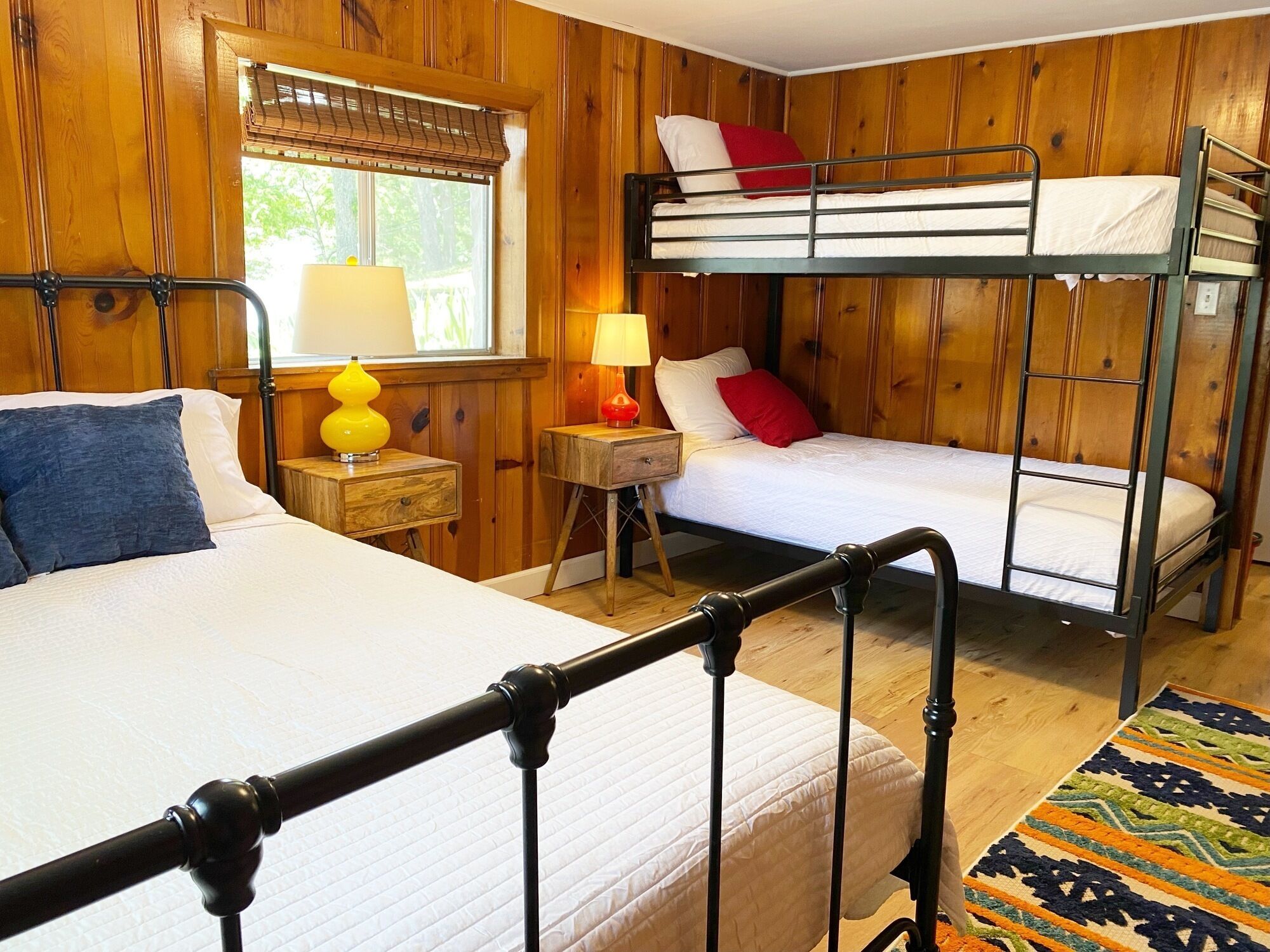 Una habitación con una cama individual, una litera, paneles de madera, una ventana con persianas, una lámpara y una alfombra.