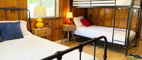 Una habitación con una cama individual, una litera, paneles de madera, una ventana con persianas, una lámpara y una alfombra.