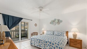 Un dormitorio con una cama, dos mesitas de noche con lámparas, un ventilador de techo, una puerta corrediza de vidrio y vistas al exterior.