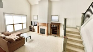 A spacious living room with a large window, a fireplace, a staircase, and a television.