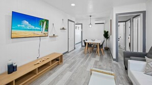 A modern living room with a flat-screen TV, a wooden bench, a dining area with a table and chairs, and a gray sofa.