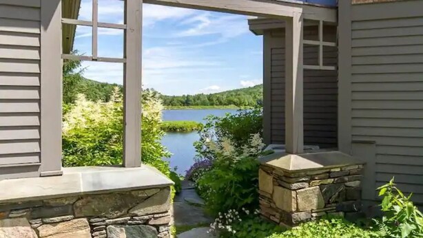 Una casa con un sendero de piedra que conduce a una terraza de madera, con vistas a un lago y un área boscosa.