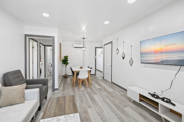 Una sala de estar moderna con un sofá, una mesa de comedor con sillas, un televisor en la pared y una planta.