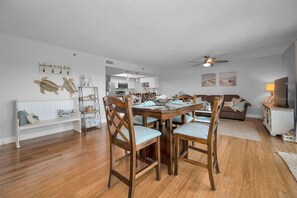 A dining area with a wooden table and chairs, a kitchen in the background, a white bench with a 'Beach' sign, and a ceiling fan.