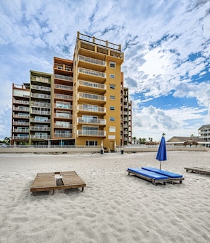 A multi-story hotel with balconies, a sandy beach in front, and a clear sky.