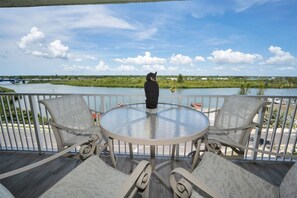 A balcony with a glass table and two chairs, overlooking a body of water and greenery.